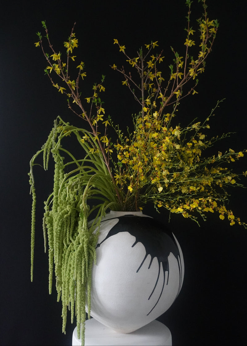 Botanical arrangement with greenery and yellow flowers in a white vase on a black background.